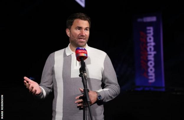 Eddie Hearn wearing a grey polo shirt with a white vertical stripe and a white blocked square