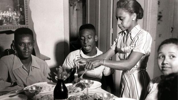 Black and white photograph of a young Pele sitting at a family meal, his father Dondinho is on his left and his mother Dona Celeste serves the food.
