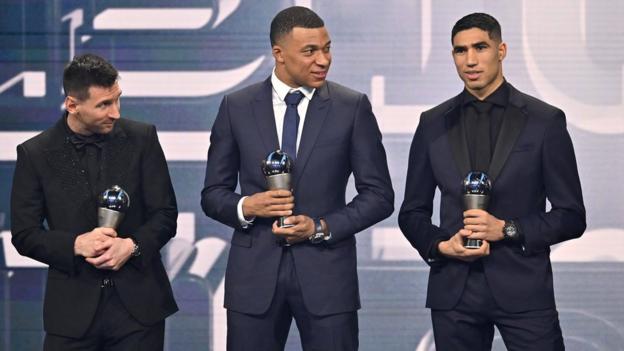 Achraf Hakimi (R) receiving his award on stage at the FIFPro awards event next to Kylian Mbappe (M) and Lionel Messi (L)