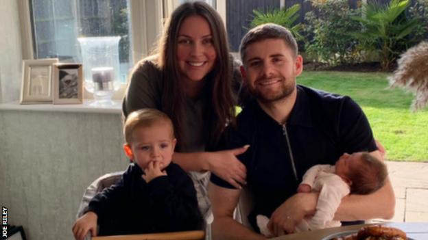 Footballer Joe Riley, his wife, Kayleigh, and children Jace (left) and Mila at home with a picture of Leo, who was stillborn, in the background
