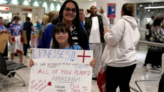 A young miss  holds up   a motion   congratulating the Lionesses, with a woman  lasting  down  her