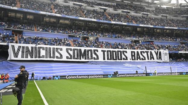 Real Madrid fans clasp  up   a banner saying “Vinicius we are all. Enough already"