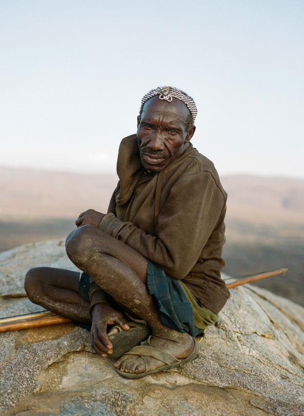 A Hadza man sits on a rocky outcrop, facing the camera with the valley below in the background