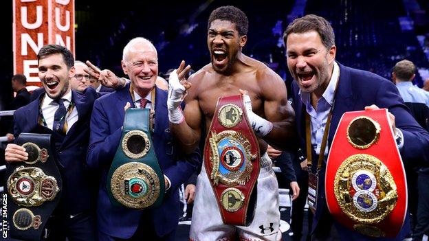 Anthony Joshua holds the belts up and celebrates with his team after beating Andy Ruiz Jr in December