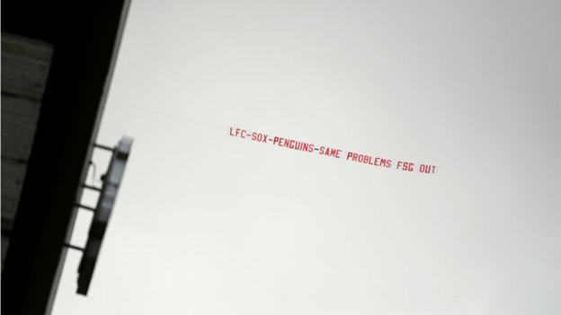 A banner is pulled across the sky over Anfield reading 'LFC - Sox - Penguins- Same problems FSG out'