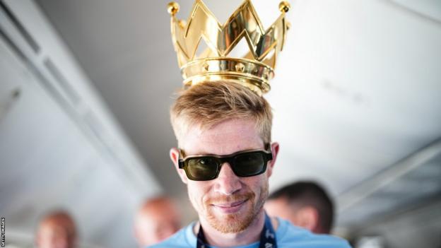 Kevin De Bruyne, sporting sunglasses wears the Champions League crown on his head as he prepares to get off the plane