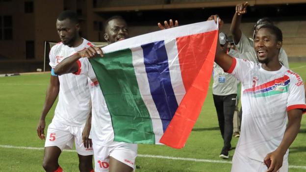Omar Colley, Musa Barrow and Alfie Jallow celebrate on the pitch with the other two players holding up a Gambian flag but Colley looking down and concentrated
