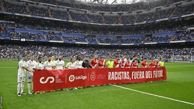 Players of Real Madrid and Rayo Vallecano enactment     up   down  a banner saying Racists, Outside of Soccer