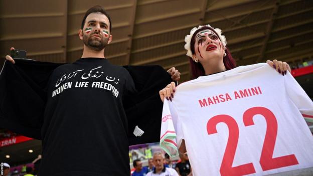 A female fan in the crowd for Wales v Iran holds up a football shirt with "Mahsa Amini - 22" printed on it and a male fan with the slogan 'women, life, freedom' on his top