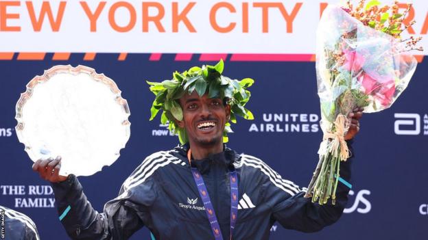 Tamirat Tola celebrates on the podium holding the title and a bouquet of flowers