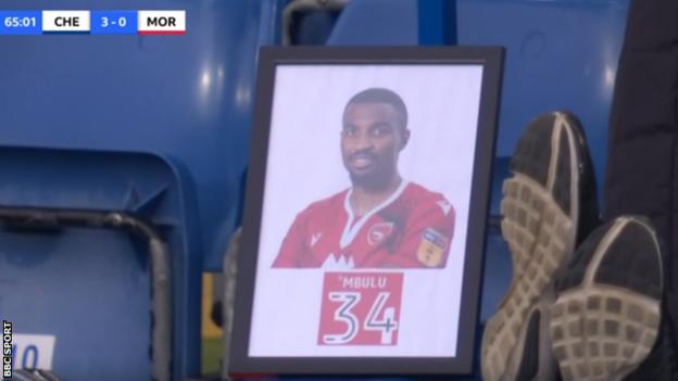 A framed picture of former Morecambe defender Christian Mbulu, who died in May 2020, on the bench at Stamford Bridge