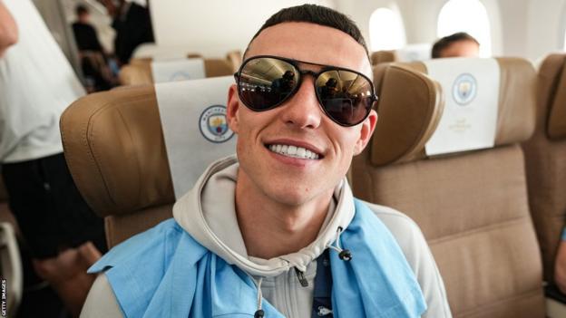 Phil Foden, wearing a pair of reflective sunglasses, grins at the camera from his plane seat