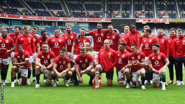 Alun Wyn Jones and the Lions squad with their trophy. Jones is wearing a tracksuit top and has his left hand in his pocket.
