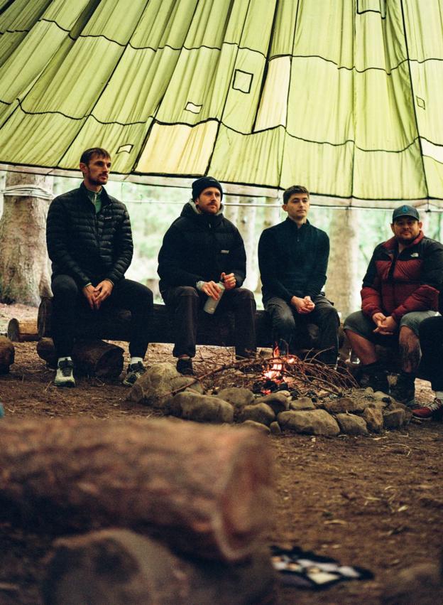 Four men sit on a log under a canopy, with a campfire in the foreground
