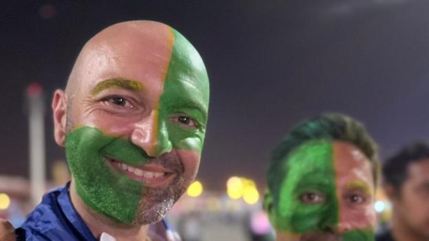 A man with half his face painted green smiles at camera