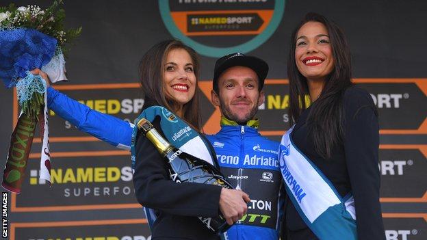 Adam Yates smiles stood between two podium girls after moving into the race lead at Tirreno-Adriatico