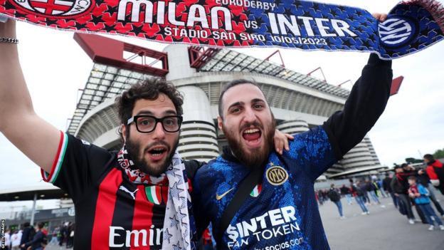 Two fans, one wearing an Inter shirt, the other a Milan shirt hold a half and half scarf aloft outside San Siro.