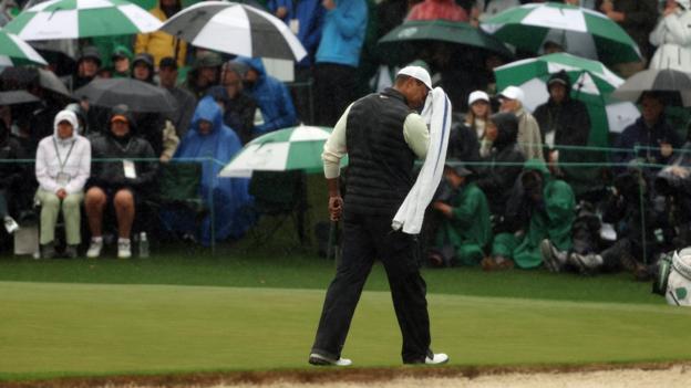 Tiger Woods wipes his face with a towel with patrons shielding themselves from the rain under umbrellas