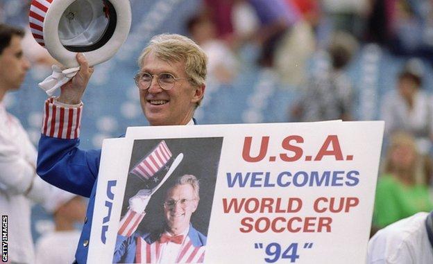 Un bărbat în uniformă cu steag american ține un banner care anunță bun venit al Statelor Unite ale Americii la cea de-a 94-a Cupă Mondială FIFA, care îl înfățișează și în aceeași uniformă purtând pălăria.