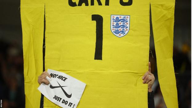 A fan holds up a cardboard cut out of a Mary Earps goalkeeper shirt with a sign calling on Nike to 'Just Do It'.