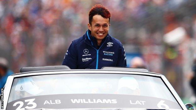 Williams driver Alex Albon smiles while being driven in a car