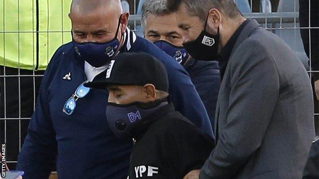 Diego Maradona wearing a DM face mask and flanked by two men