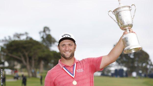 Jon Rahm holds up the US Open trophy and smiles with a medal round his neck
