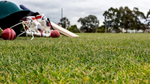 A cricket helmet, bat, gloves and two cricket balls on the outfield of a cricket pitch