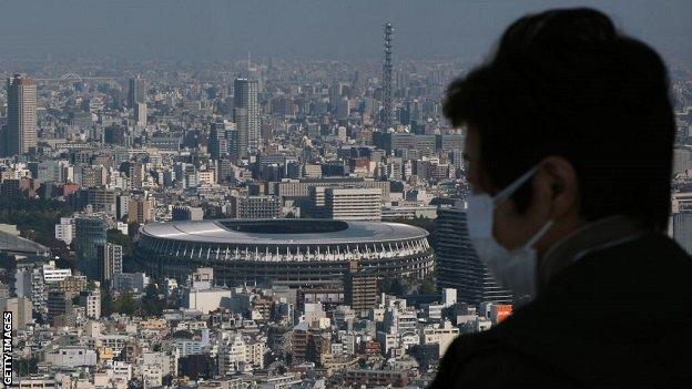 A masked spectator overlooks the Tokyo skyline, which features the Olympic Stadium