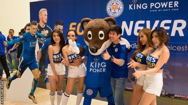 A mascot, models and a fan pose for a photo at King Power HQ